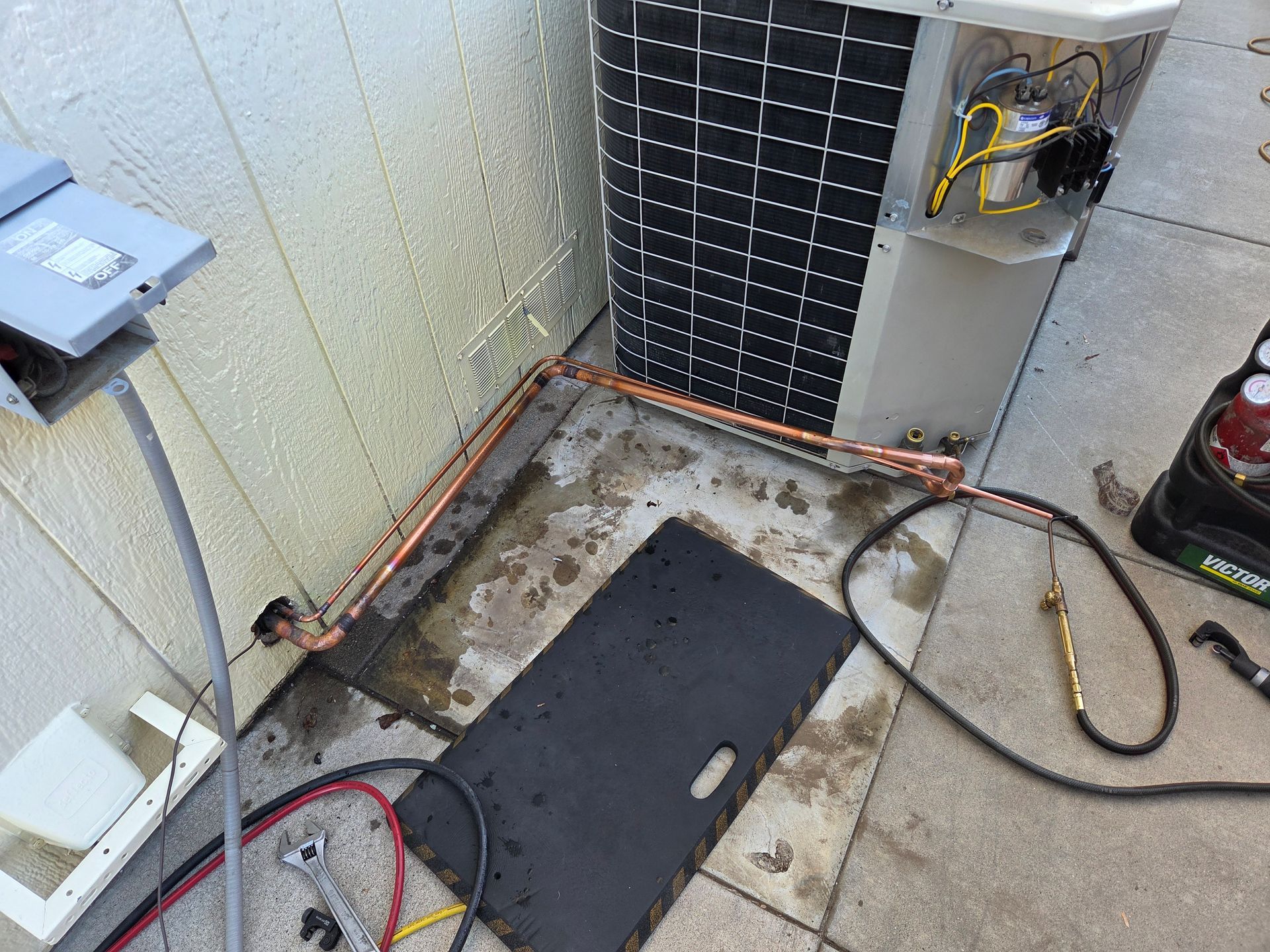 HVAC unit installation outdoors; copper tubing, tools, and electrical box on a concrete patio.