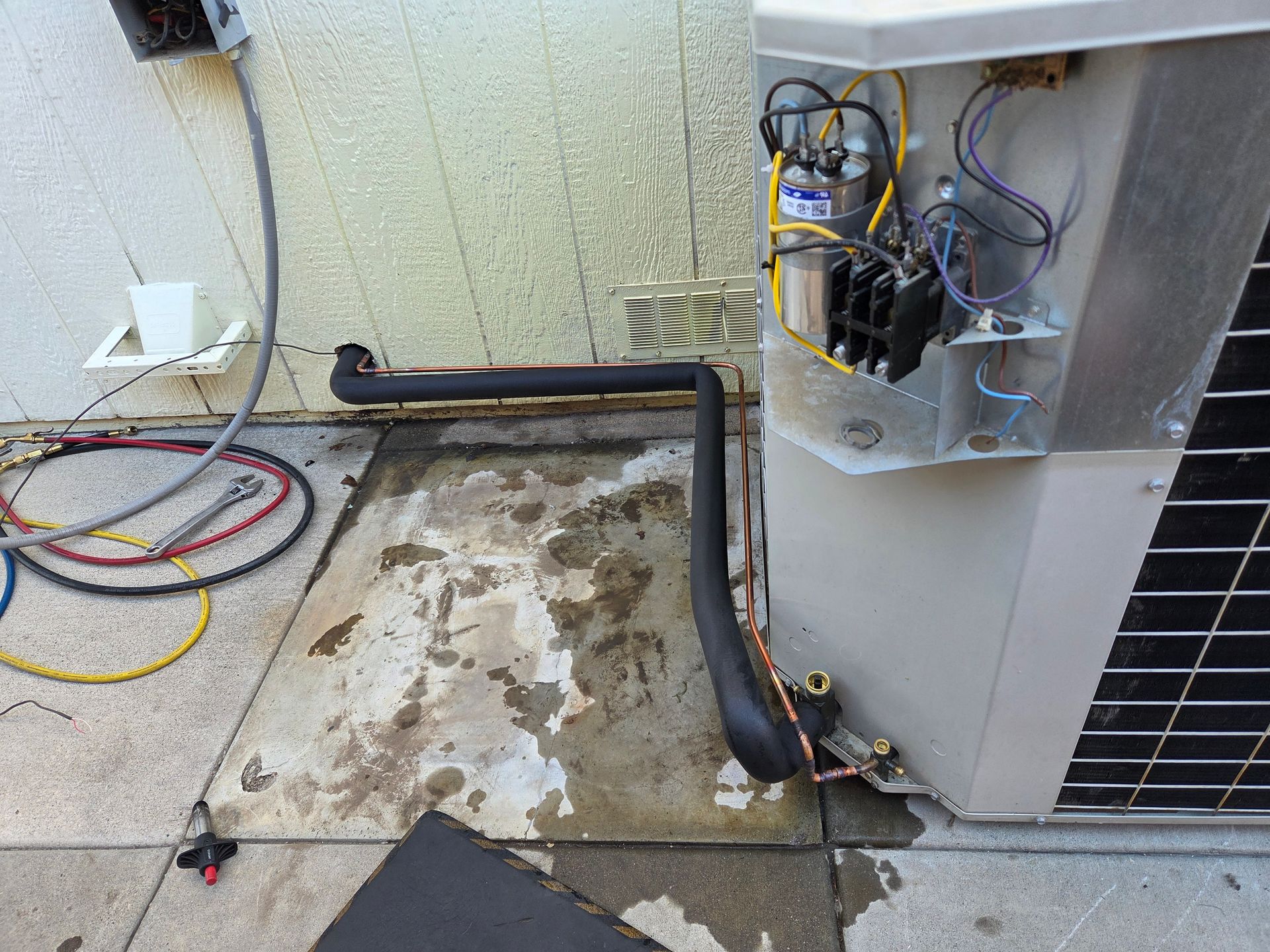 HVAC unit with exposed components. Copper pipes with insulation on a wet patio. Electrical wiring visible.