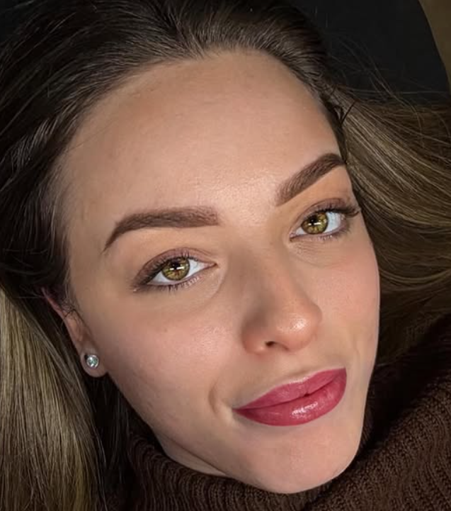 Primo piano di una donna con rossetto lucido e sopracciglia ben definite, che guarda in camera su uno sfondo scuro.