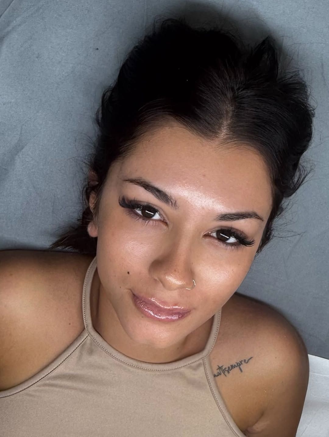 Primo piano di una persona sdraiata su una superficie grigia, che indossa una maglietta beige e guarda in camera con un'espressione dolce.