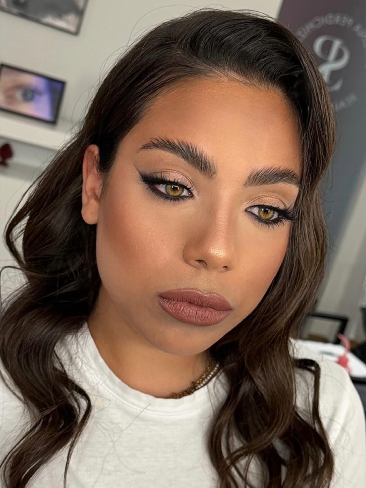 Primo piano di una persona con eyeliner alato e rossetto nude, che guarda verso il basso in una stanza illuminata da una luce soffusa.