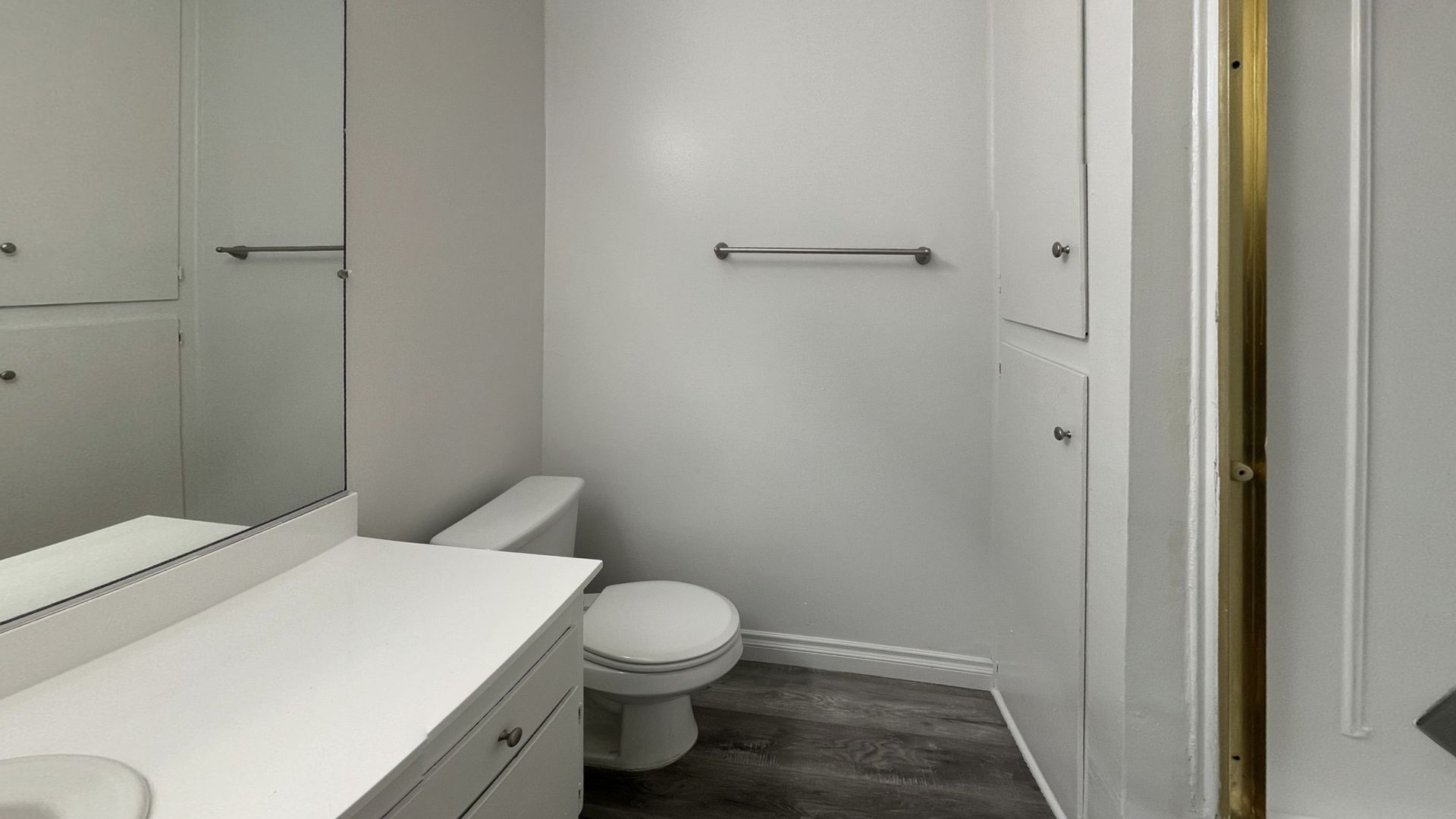 Bathroom with a white vanity, toilet, cabinets, and shower stall with gold trim; gray walls and flooring.