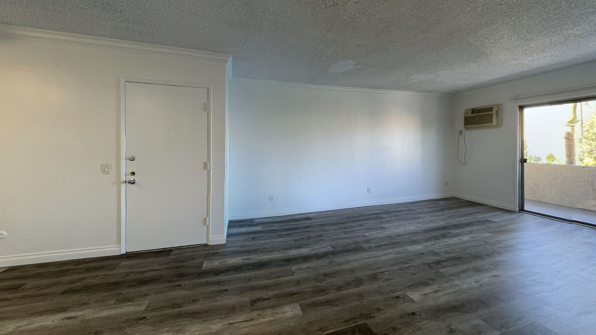 Empty apartment interior with white walls, gray wood-look flooring, door, sliding glass door to balcony.