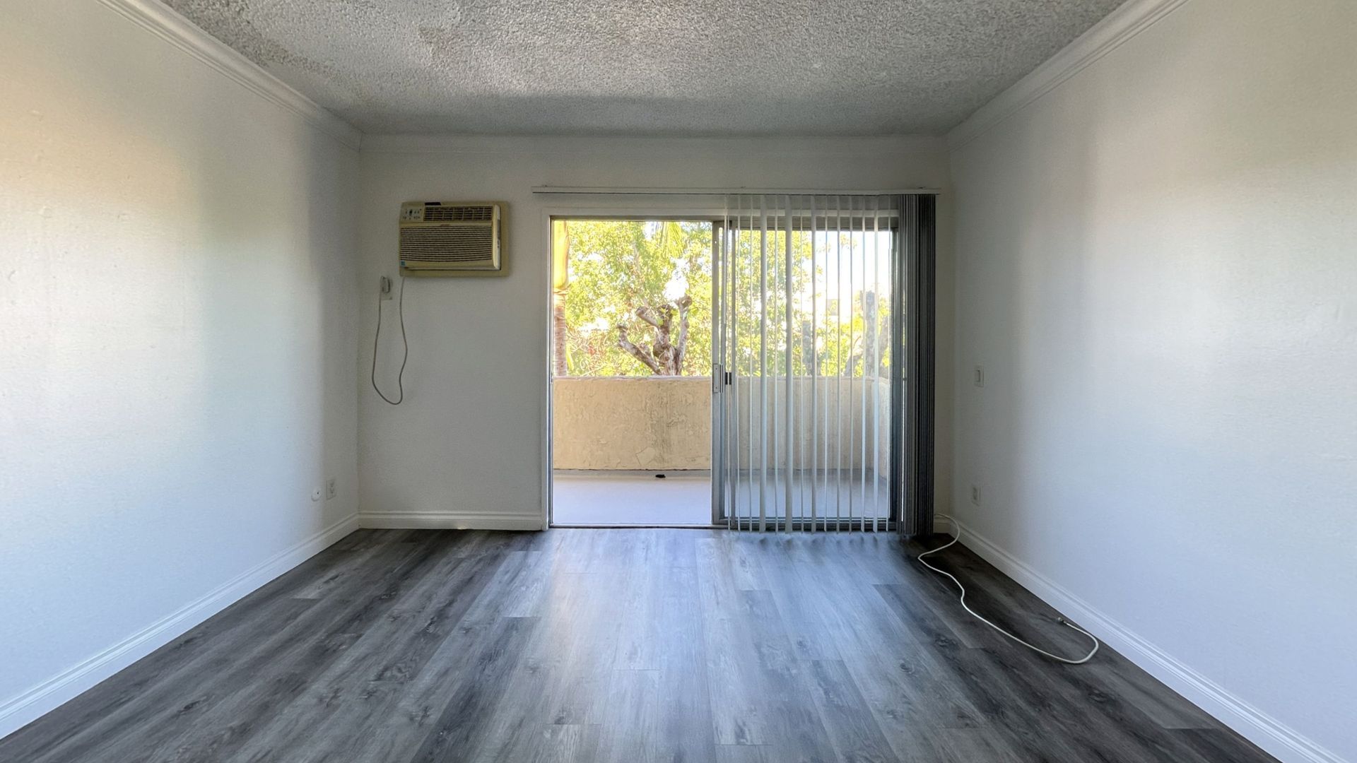 Empty room with sliding glass door leading to balcony, gray flooring, white walls, AC unit.
