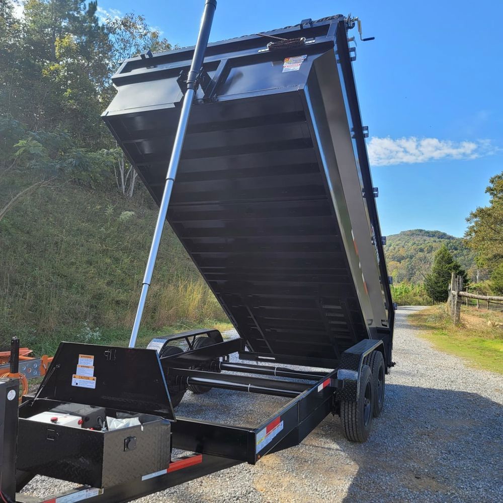 Black dump trailer with bed raised in front of green foliage and a wooden fence.
