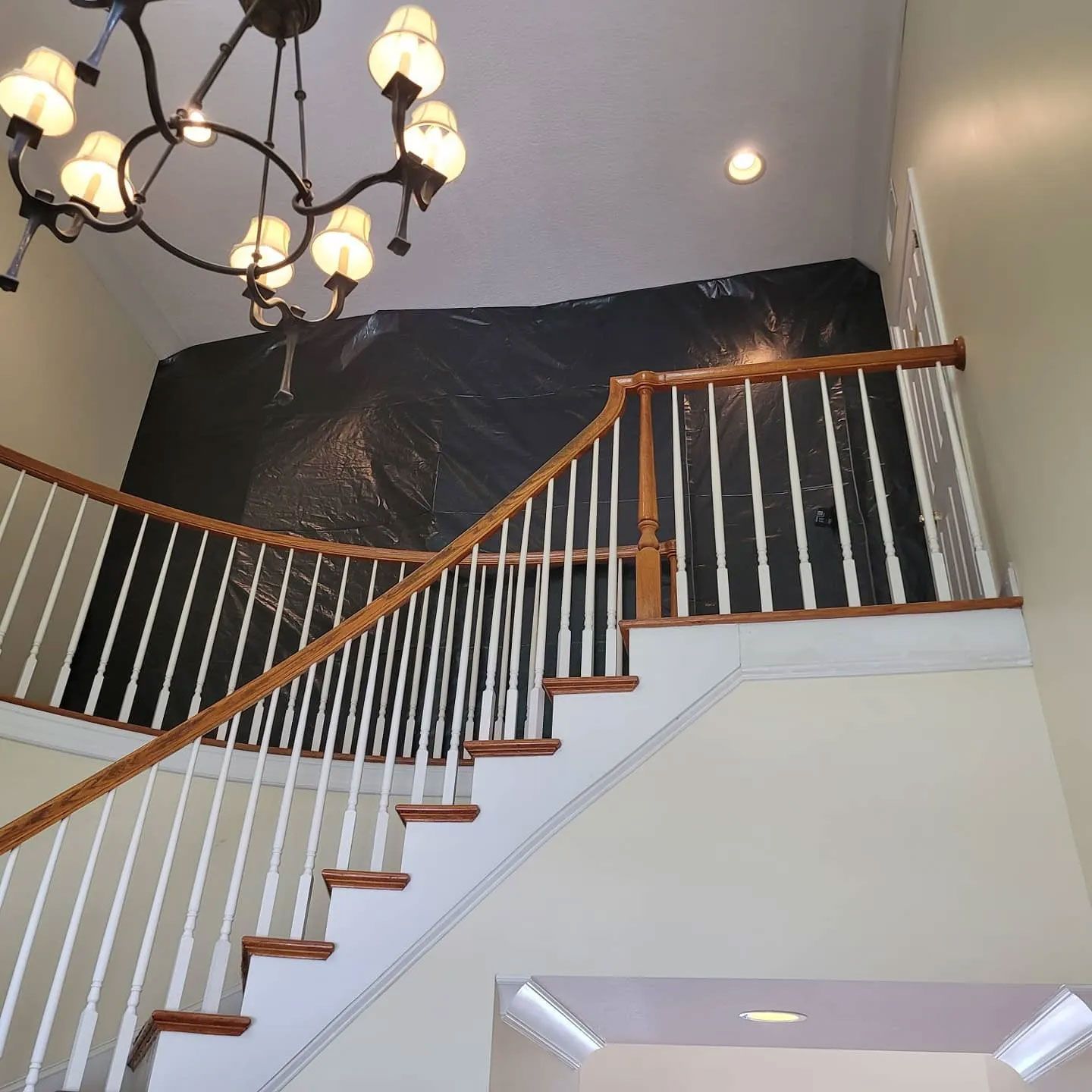 Staircase with wooden handrail, leading up to a black tarp on a high wall, and a chandelier.