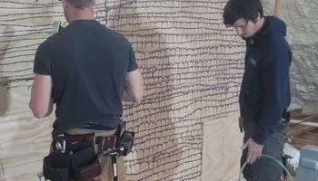 Two construction workers applying sealant to plywood wall. One wears a tool belt.