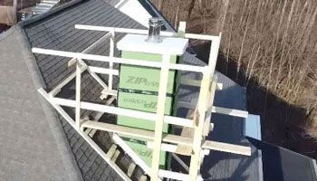 Wooden scaffolding surrounds a chimney on a dark roof. Green insulation is visible on the chimney.