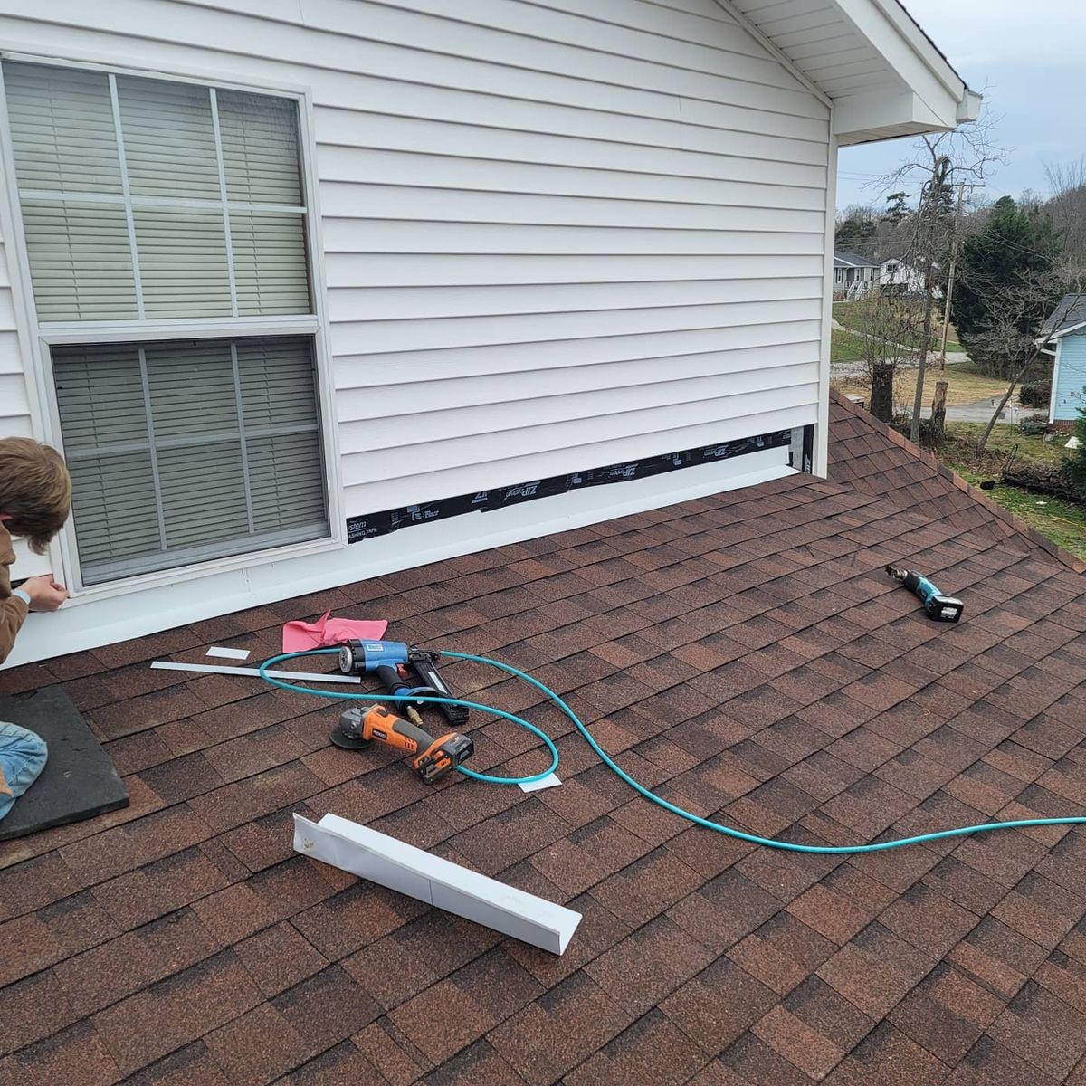 A person works on a roof near a window. Tools and materials are scattered on the shingles.