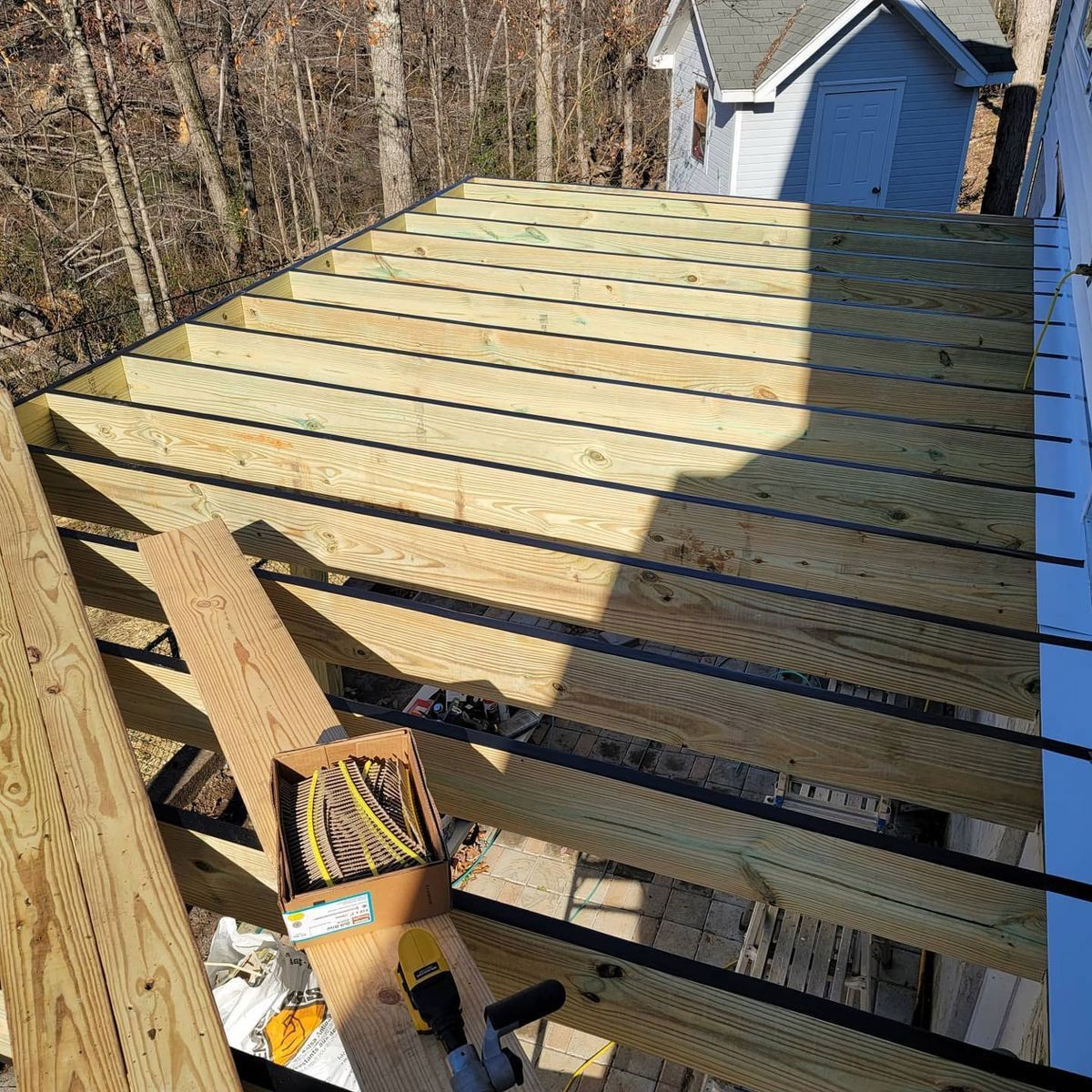 Wooden deck under construction.  Lumber beams, partly assembled, box of wires visible.  Sunlit outdoor setting.