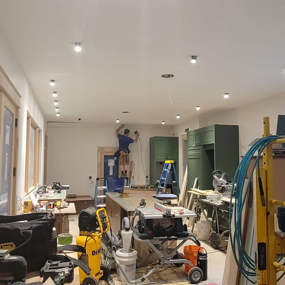 A man on a ladder installs ceiling lights in a cluttered workshop. Tools and equipment are everywhere.