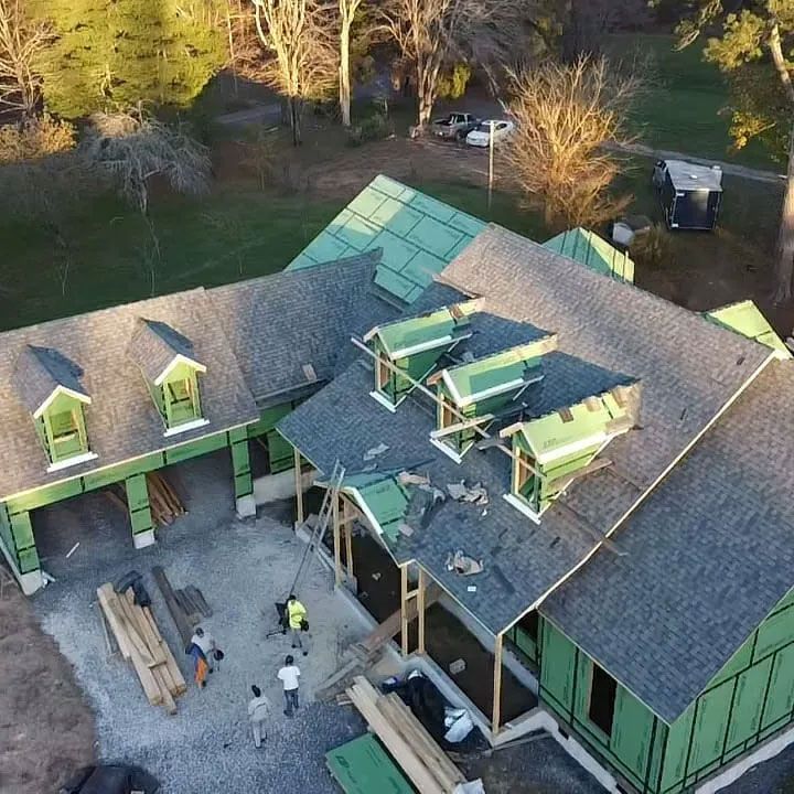 Construction of a house with workers on the roof and on the ground. Green wrapping, wood framing, and grey roof.