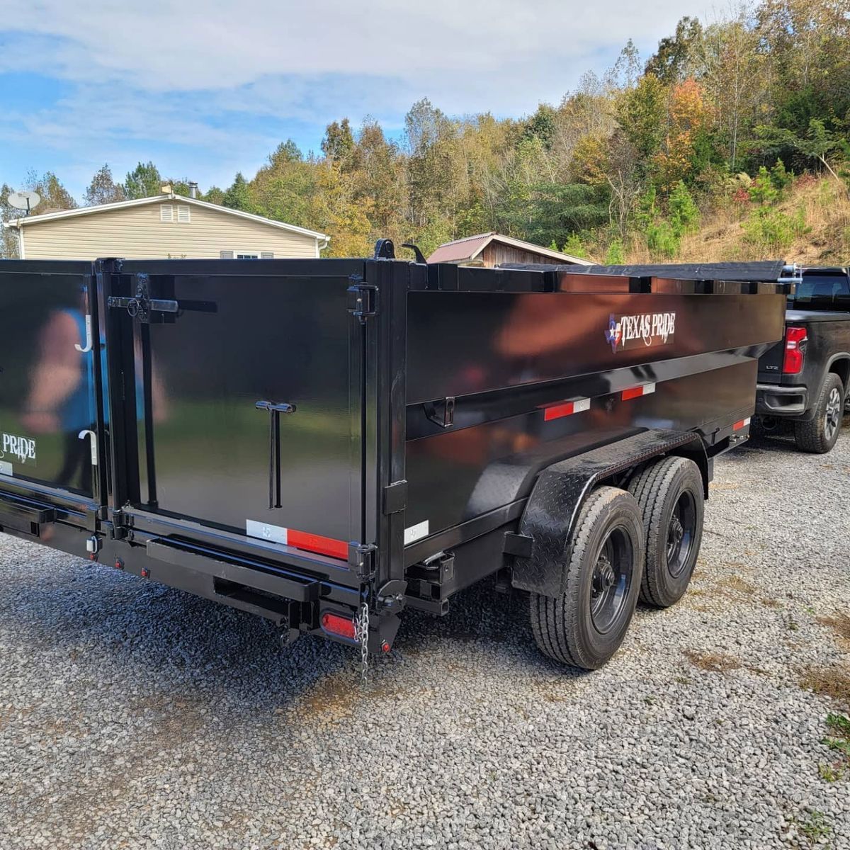 Black dump trailer hooked to a truck on a gravel driveway, trees and a blue sky in the background.