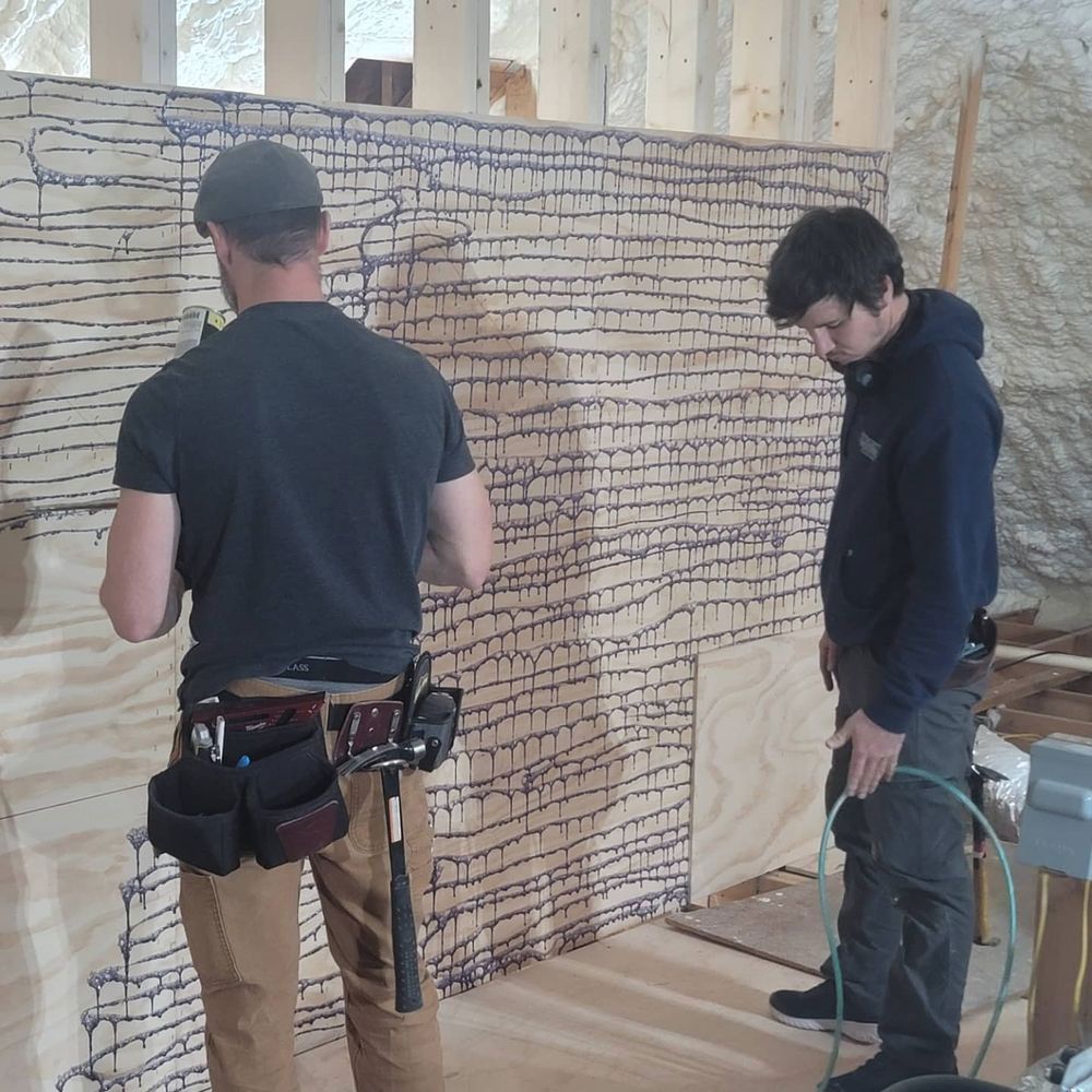 Two men working on a wooden wall marked with blue lines. One in a black shirt, the other in a blue hoodie. Interior setting.
