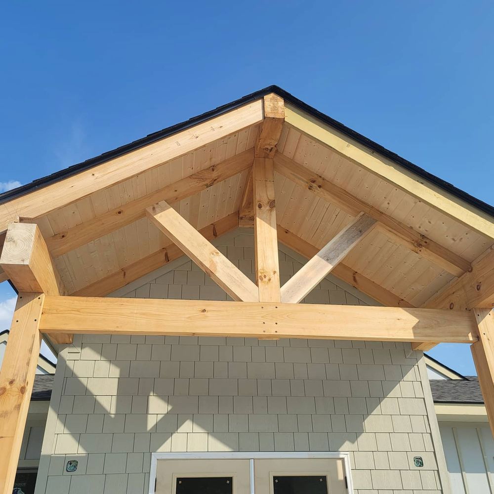 Wooden porch roof with exposed beams, over a building with light green siding, against a blue sky.