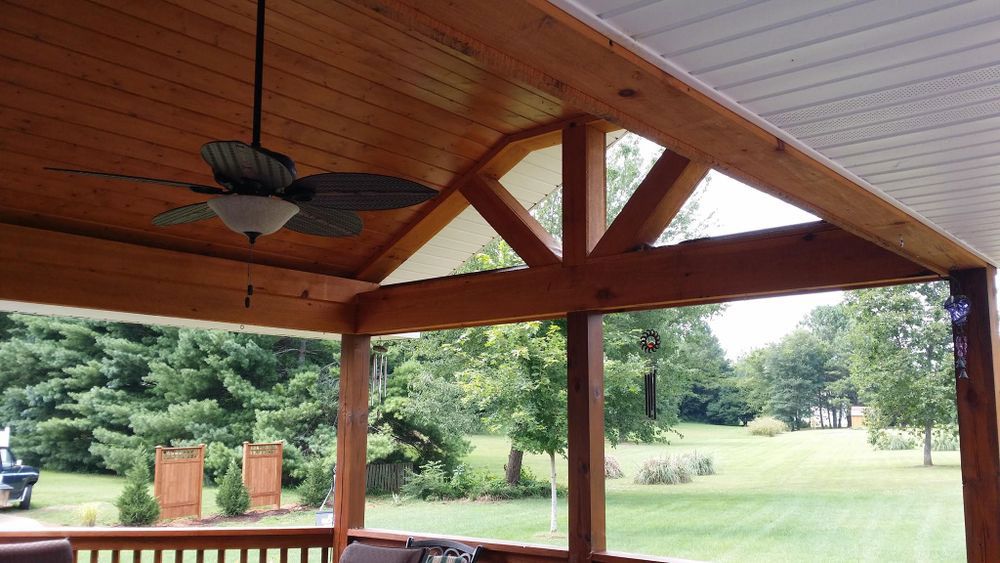 Covered porch with wooden beams and ceiling fan, overlooking a green yard.