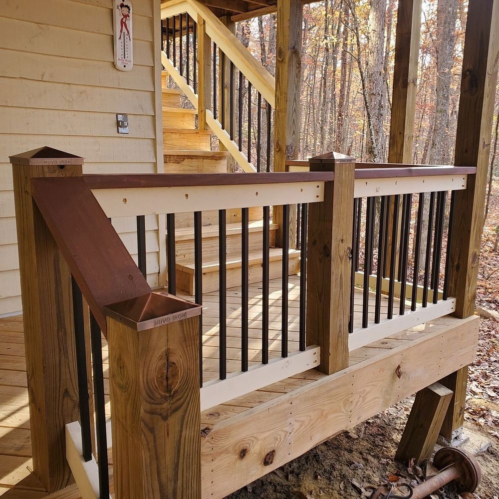 Wooden deck railing with black spindles, leading to stairs, surrounded by trees.
