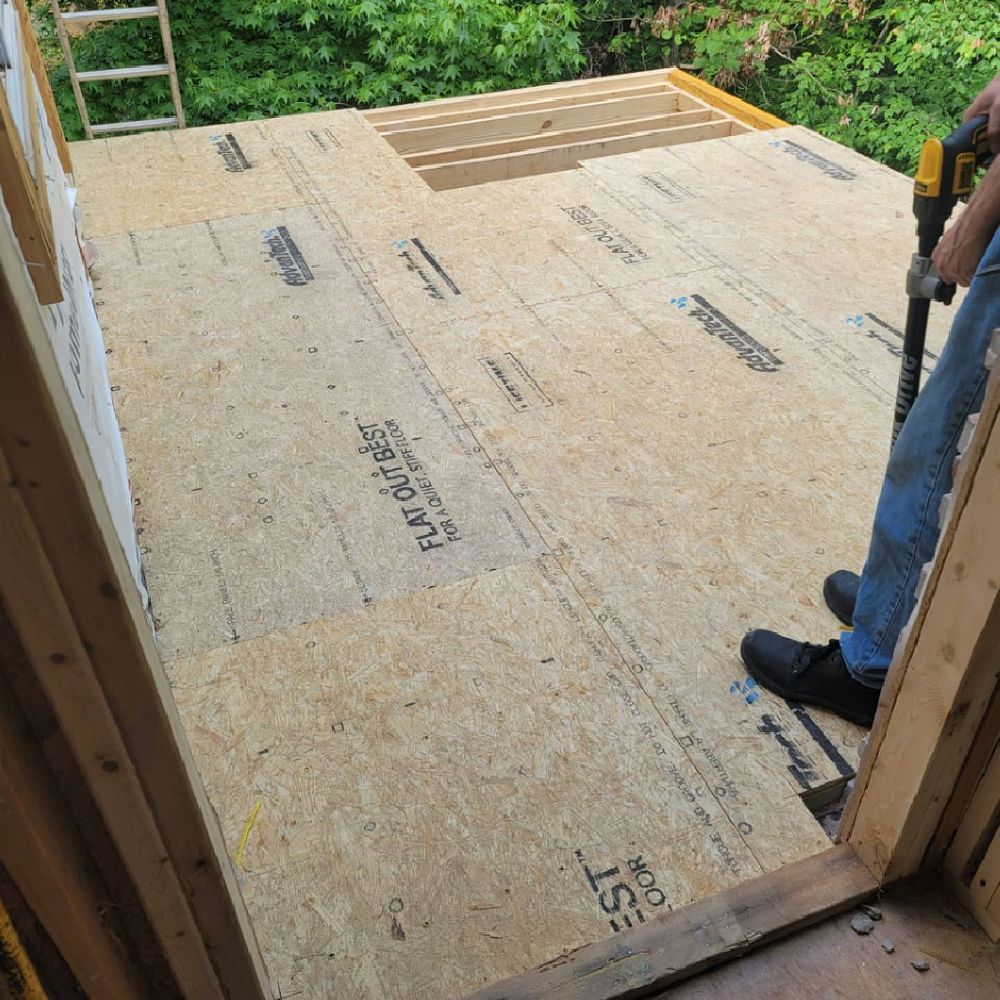 Person using a drill to install flooring in a wooden structure. Light-colored wood with cut-out opening.