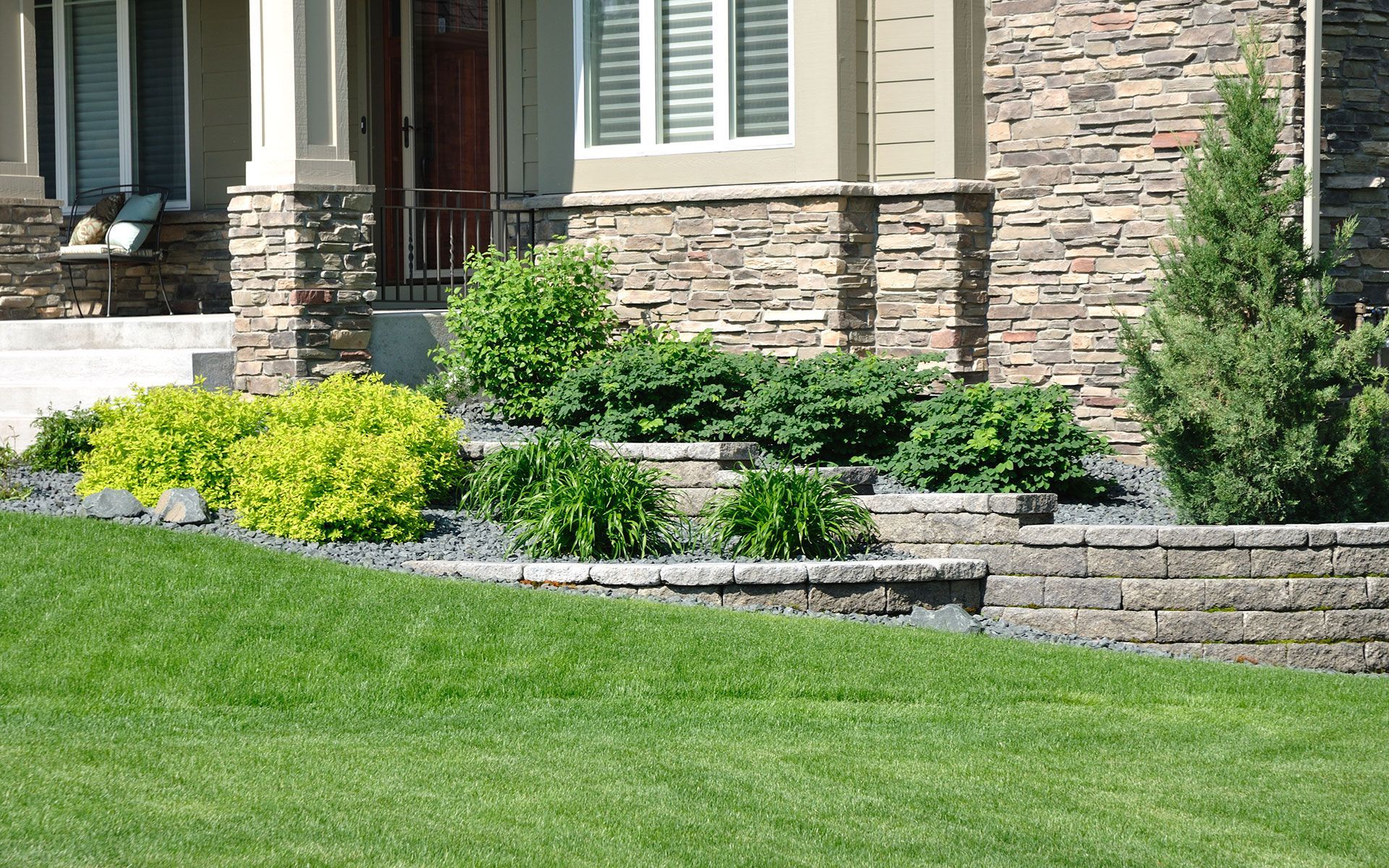 Lush front yard with tiered stone wall, various green and yellow plants, and a house with stone veneer.