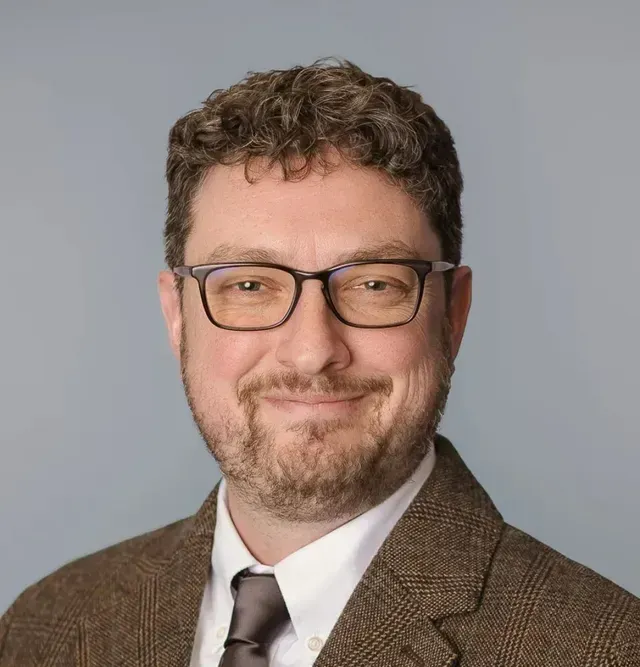 A man wearing glasses and a suit and tie is smiling for the camera.