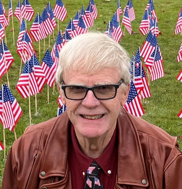 A bald man wearing a red jacket is smiling in front of a plant.