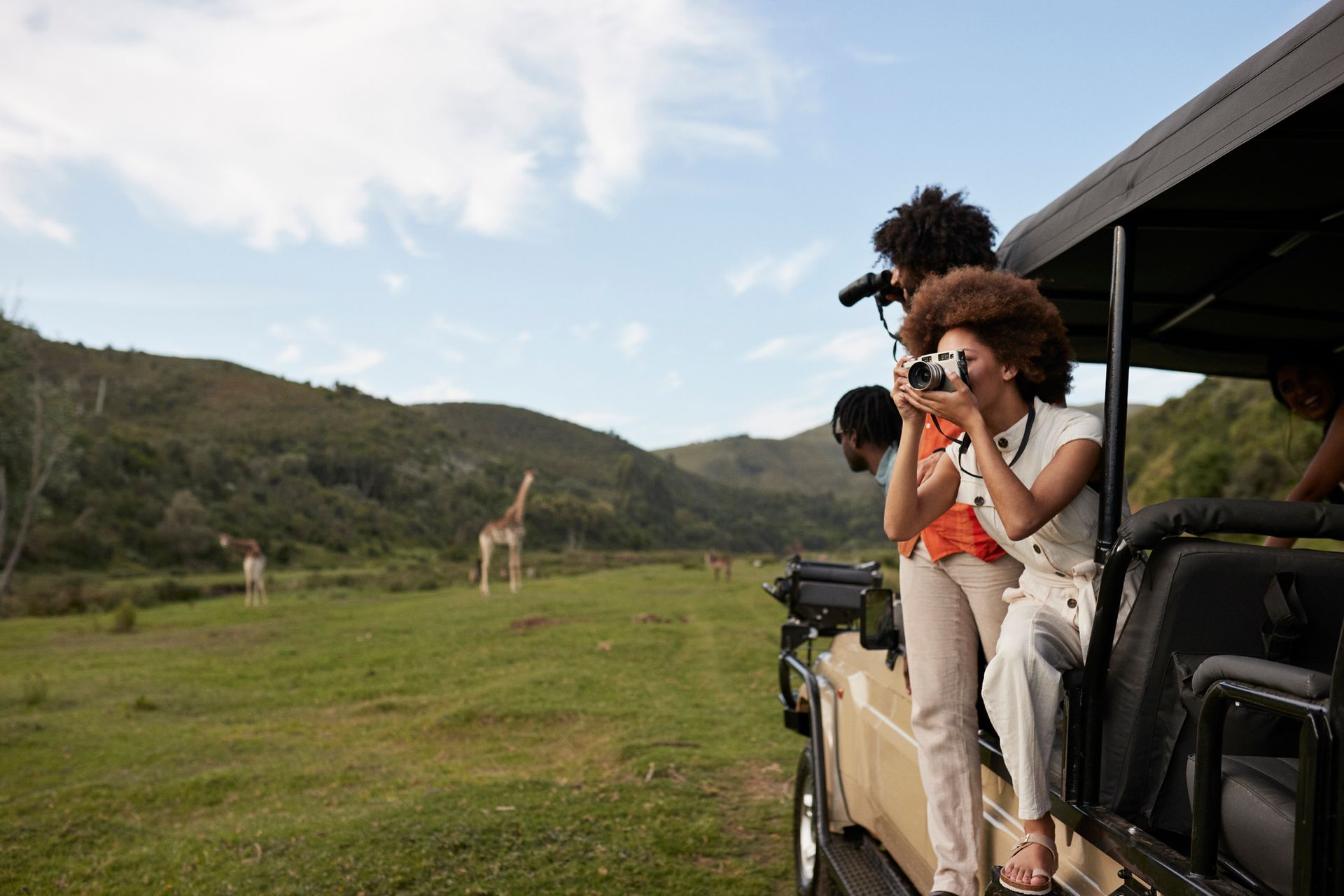 People on safari in open-top jeep, photographing giraffes in green grassy field.