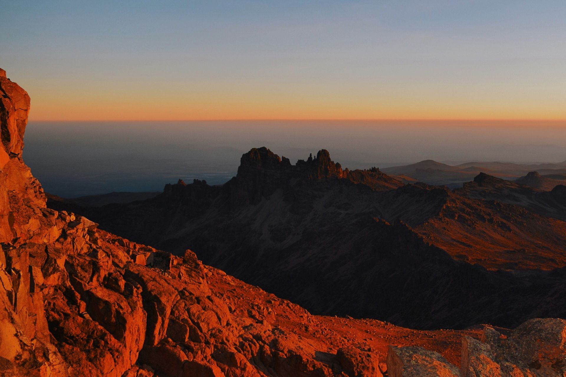 Sunrise over jagged, rocky mountain peaks with golden and orange hues, clear blue sky.