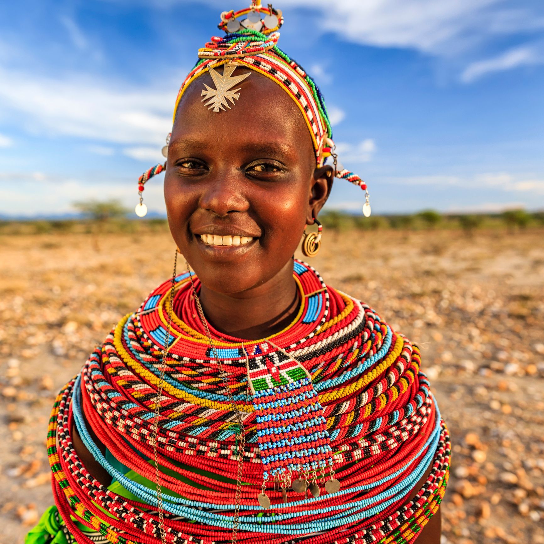 A smiling Maasai woman in traditional colorful beaded jewelry and headwear in a sunny, arid landscape.
