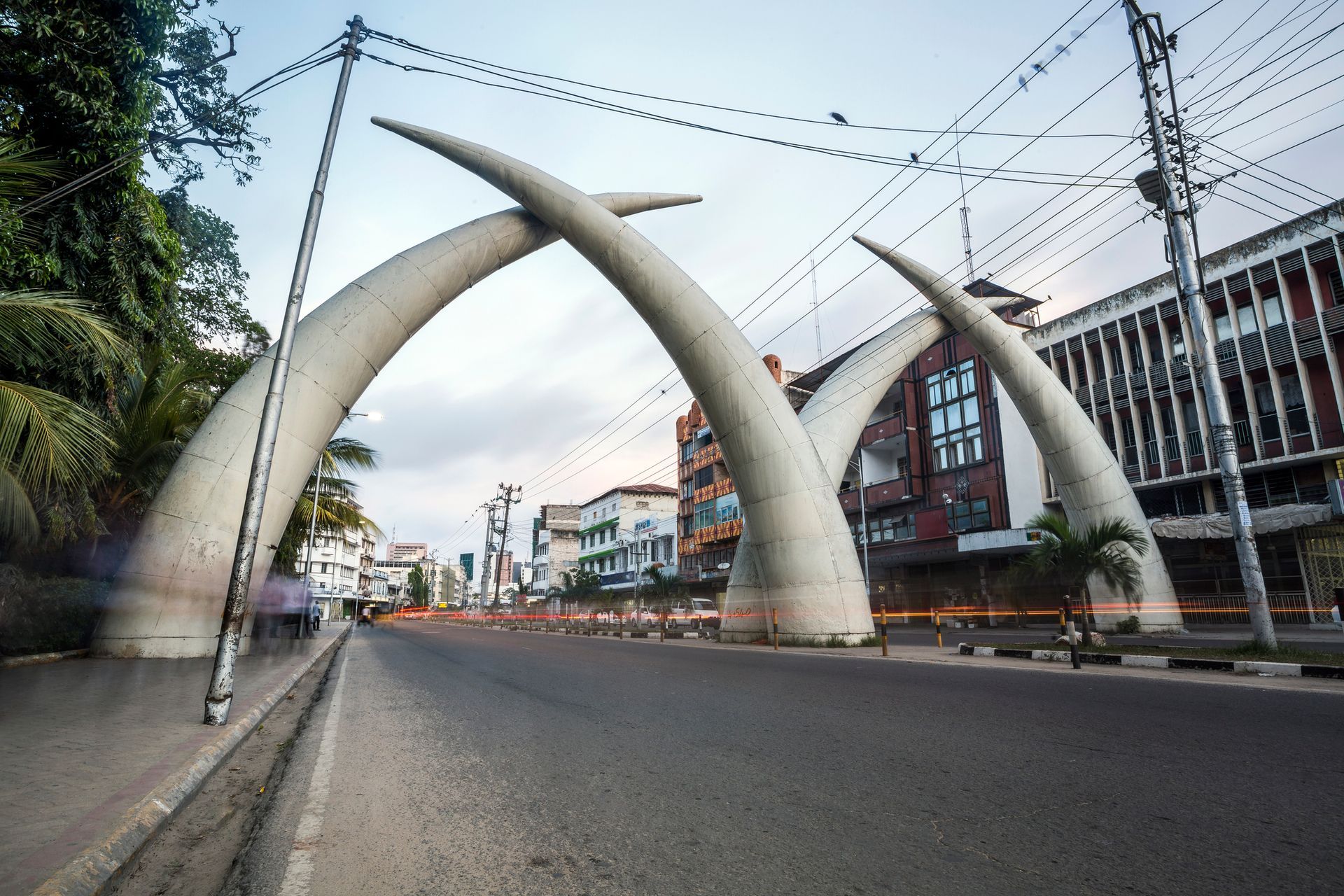 Giant ivory-colored tusks arched over a road in Mombasa, Kenya, with buildings and traffic visible.