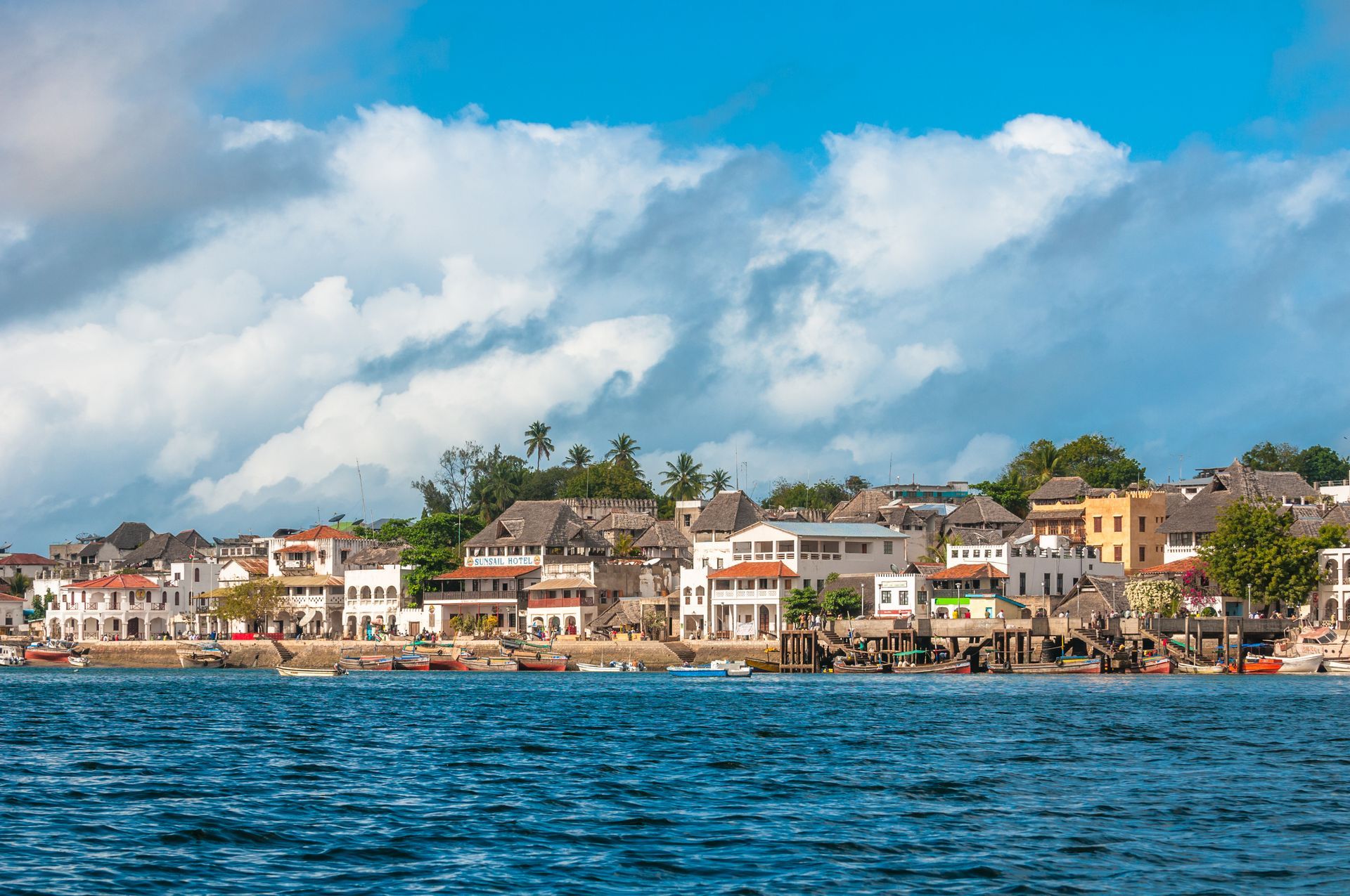 Coastal town with white buildings and thatched roofs, viewed from the water under a cloudy blue sky.