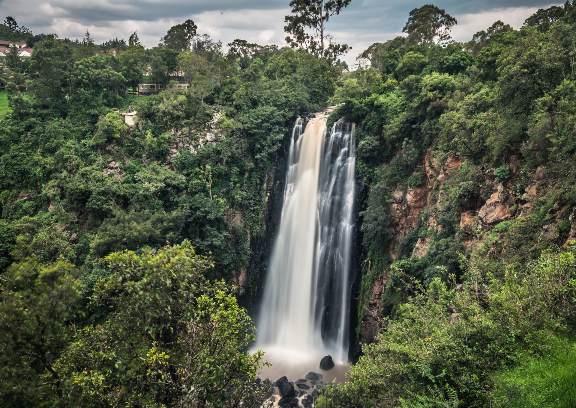 Waterfall cascading down rock face surrounded by lush green trees under a cloudy sky.