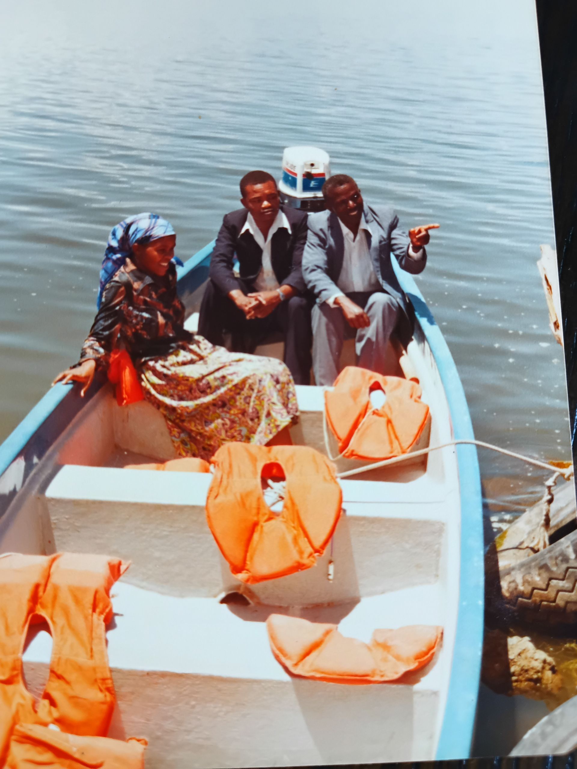 Three people in a blue boat on water; one woman wearing a headscarf, two men in suits, and orange life vests.
