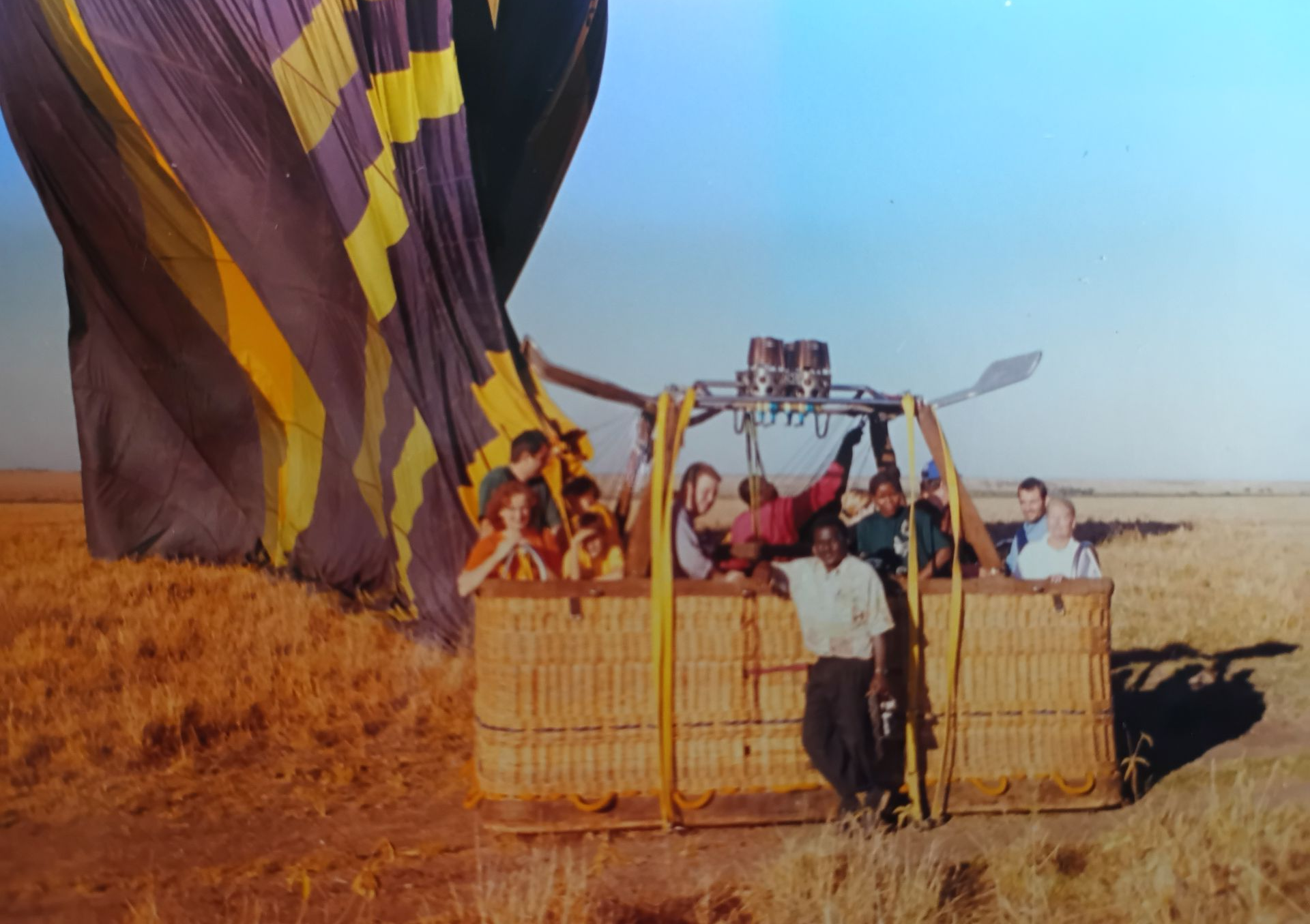 People in a hot air balloon basket preparing for takeoff in a field. Balloon is purple and yellow.