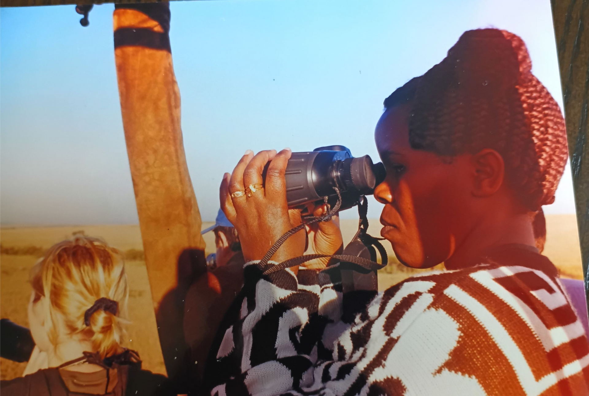 Woman looking through binoculars, wearing patterned sweater, near a wooden support in a sunny, outdoor setting.