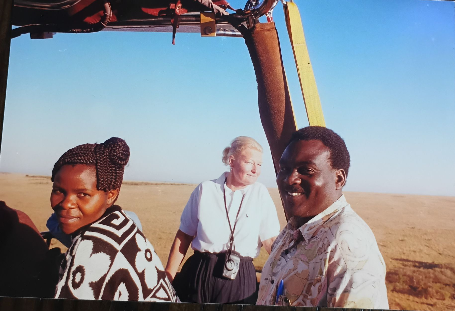 Three people in a hot air balloon over a savanna. Smiling, they are looking at the camera.