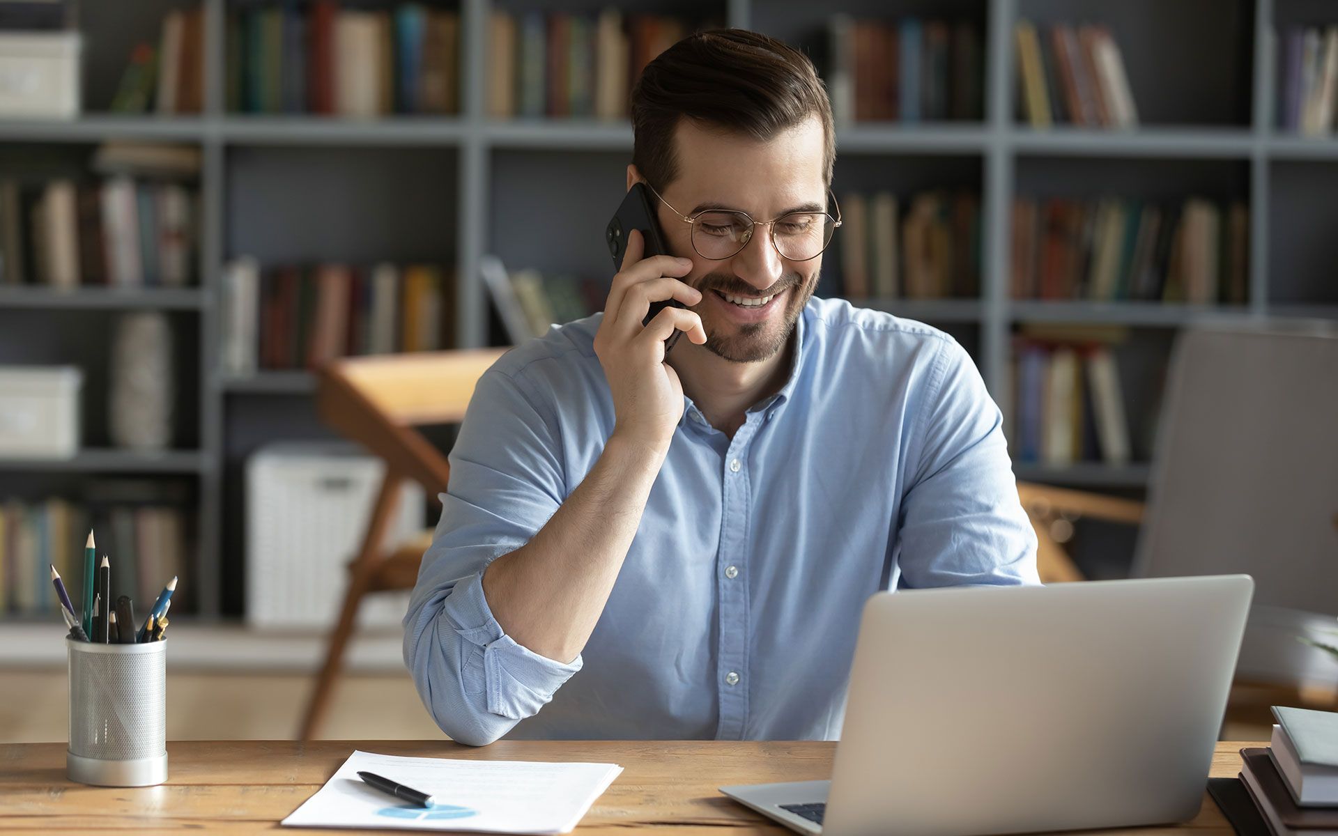 Man smiles while on phone at a desk with a laptop in front of a bookcase.