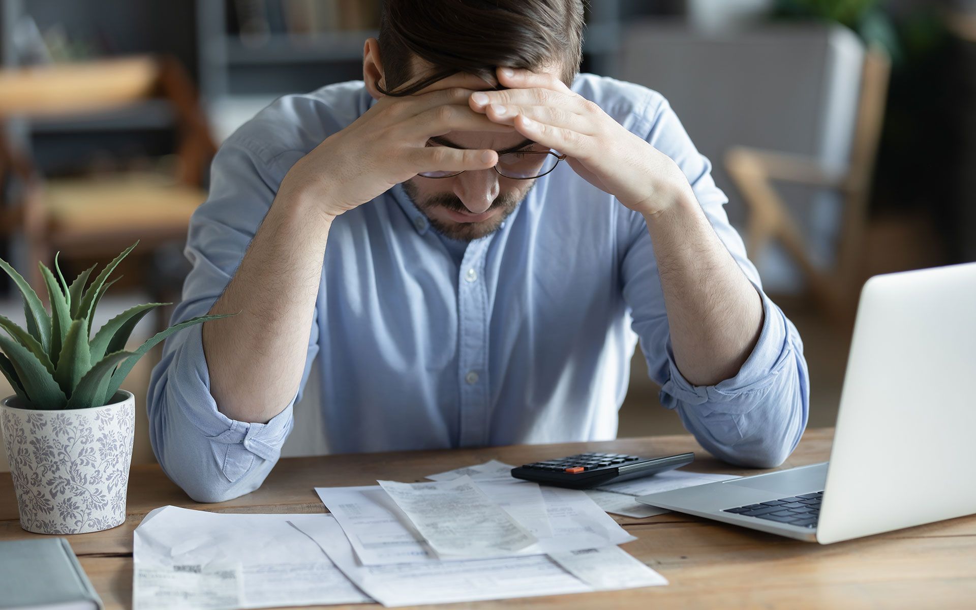 Man with head in hands, looking stressed, at desk with bills, calculator, and laptop.