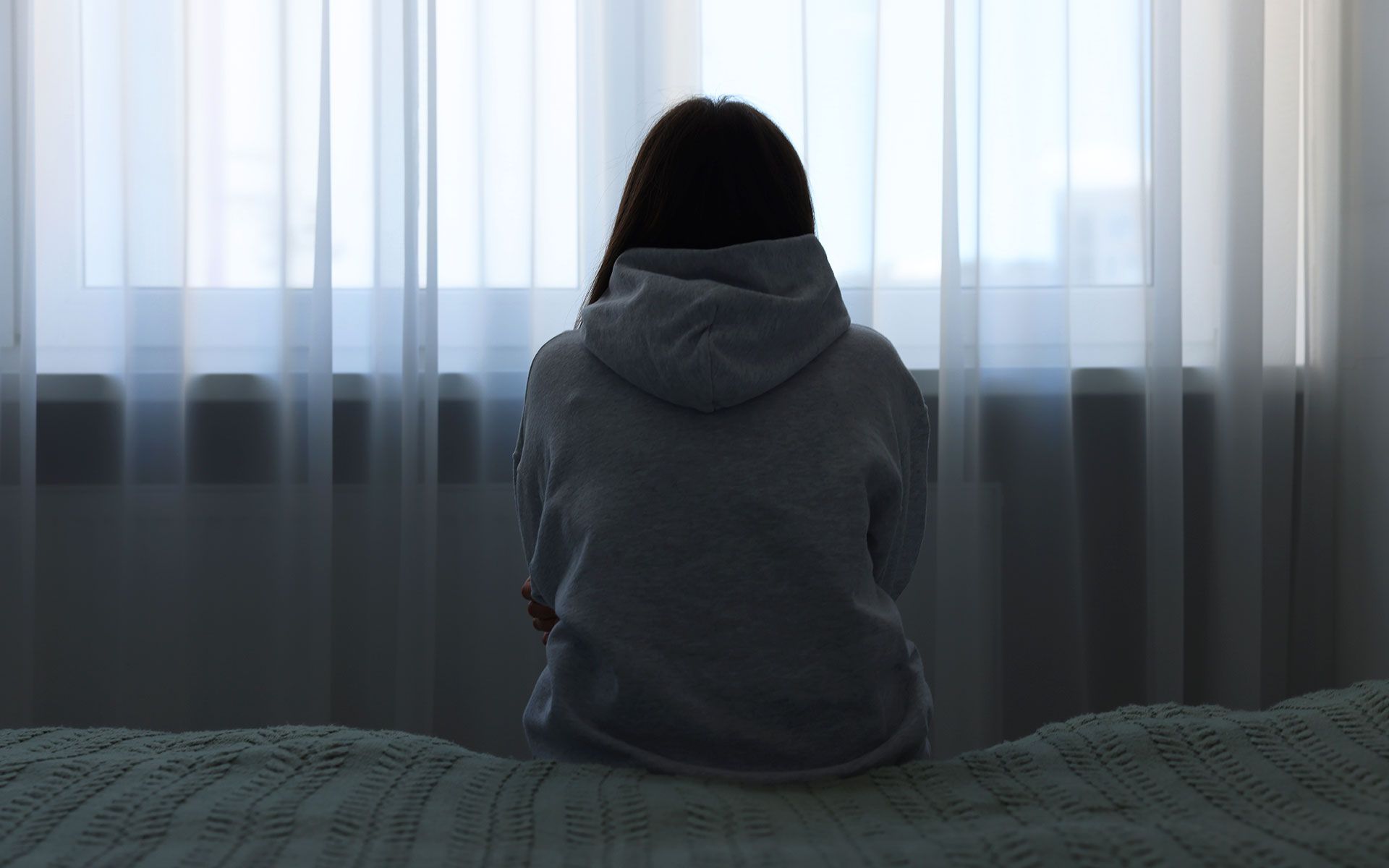 Woman in hoodie sits on bed, looking out a window with sheer curtains.