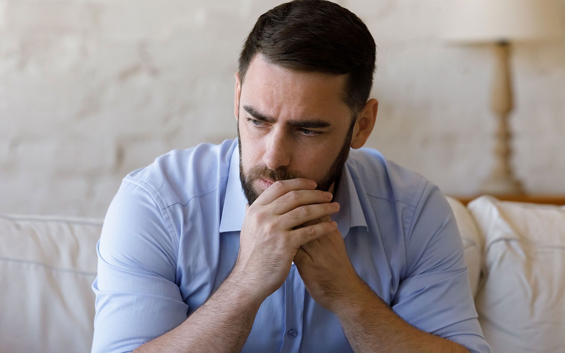 Man with a beard in a blue shirt looks down, hands clasped, appearing worried on a couch.