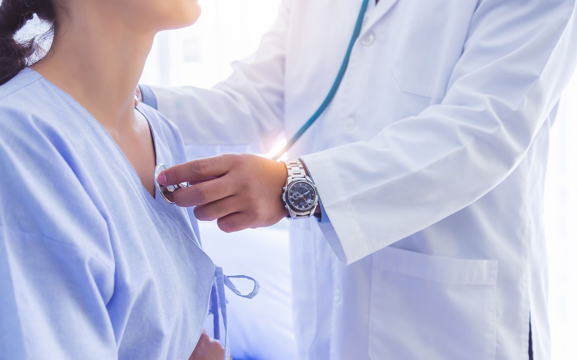 Doctor examining a patient's chest with a stethoscope; both are inside a brightly lit room.