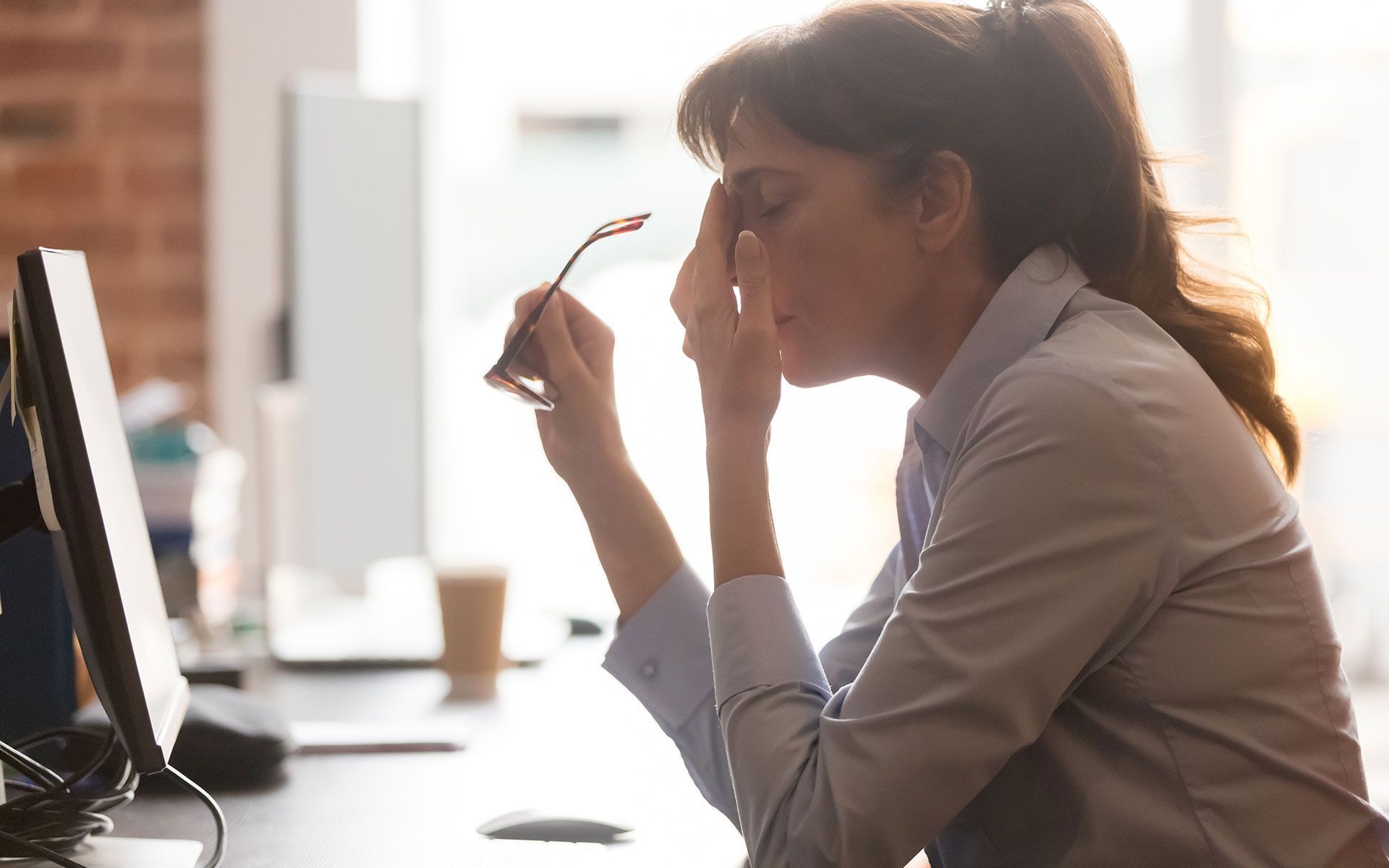 Woman at desk with eyes closed, holding glasses, looking tired.