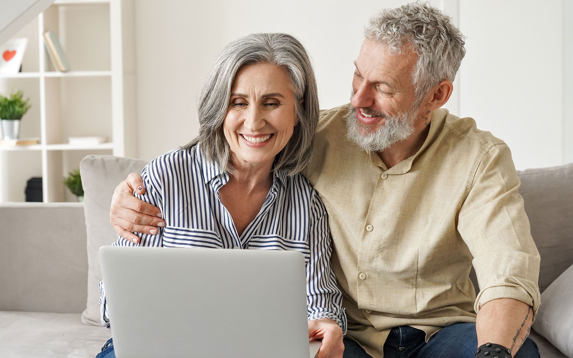 Older couple smiles looking at laptop on a couch; woman has a striped shirt, man has his arm around her.