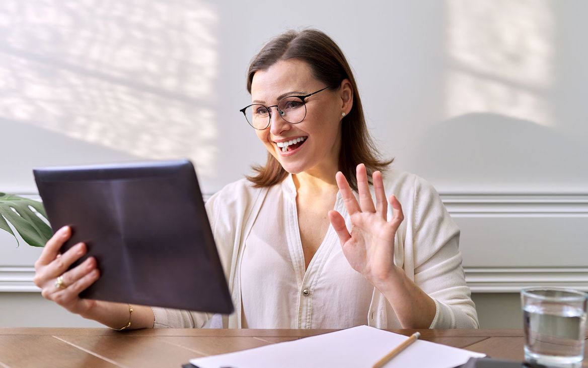 Woman with glasses waves at tablet screen, smiling. Sitting at a table, indoor setting.