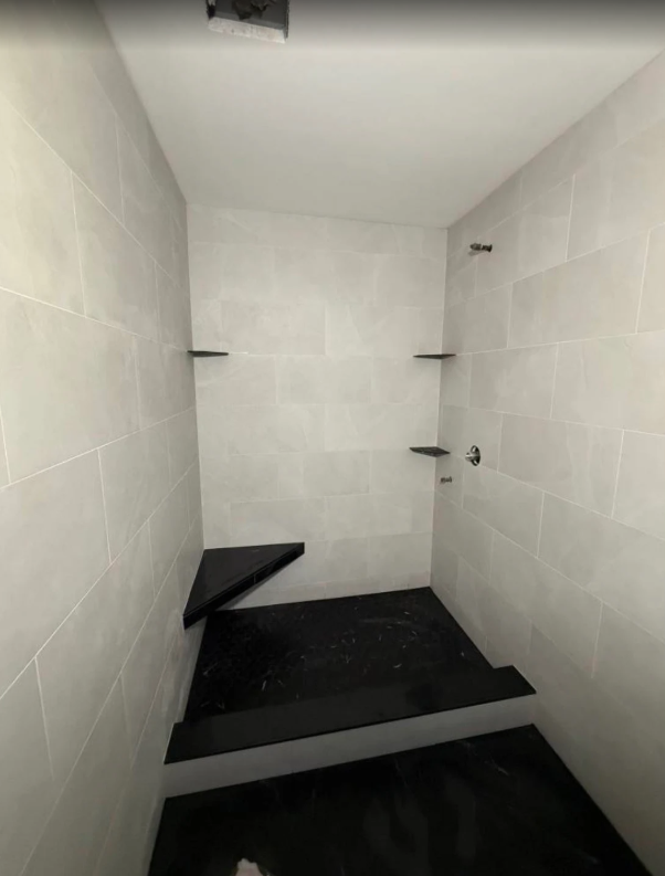 Narrow white tiled shower stall with black shower floor and small corner shelves