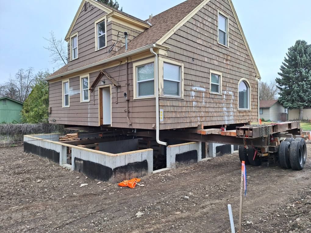 A brown house being moved on a heavy-duty flatbed trailer over a newly constructed concrete block foundation.