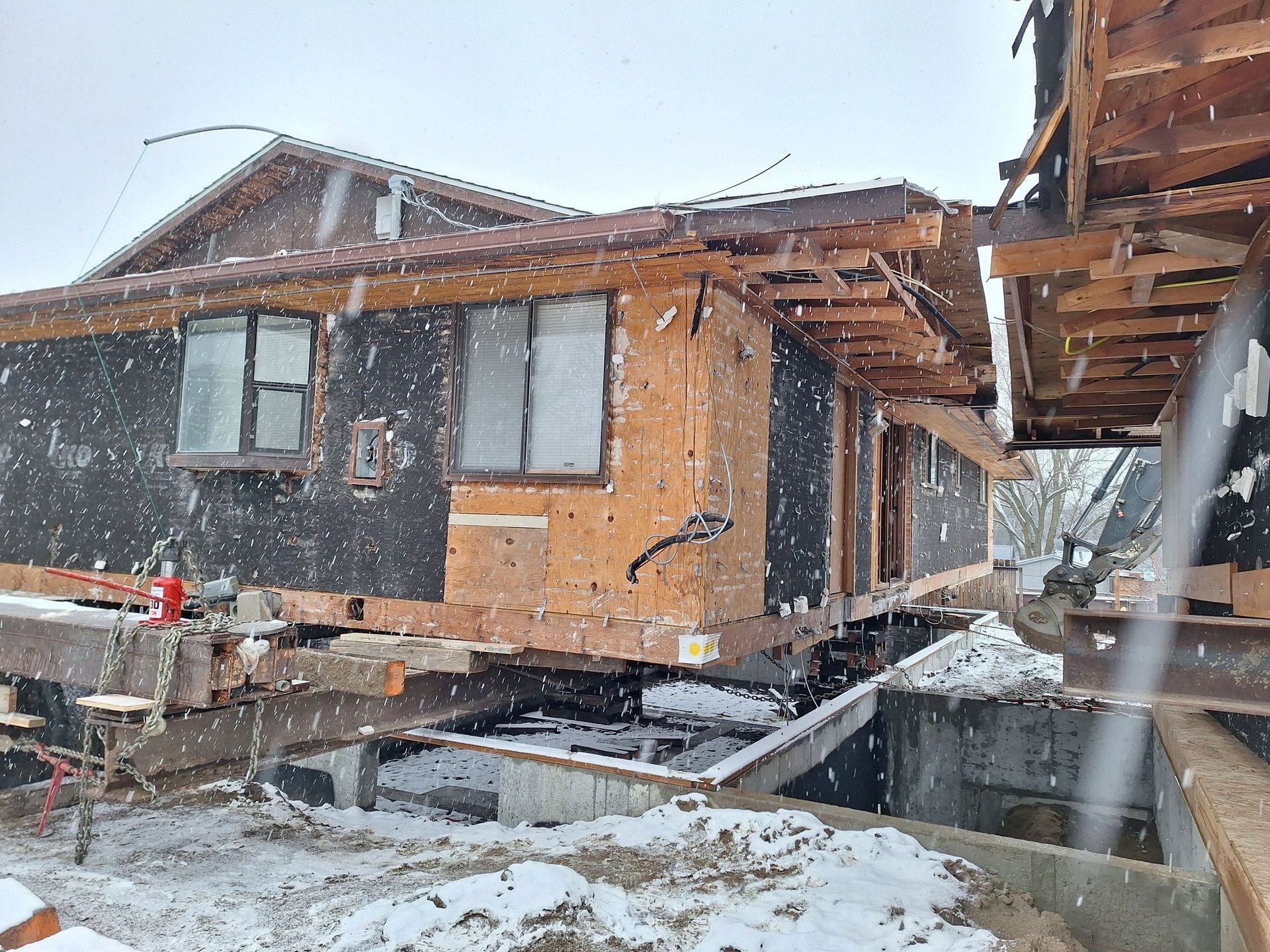 A house during a snowy day, elevated on steel beams for foundation work, with exposed framing and dark exterior paper.