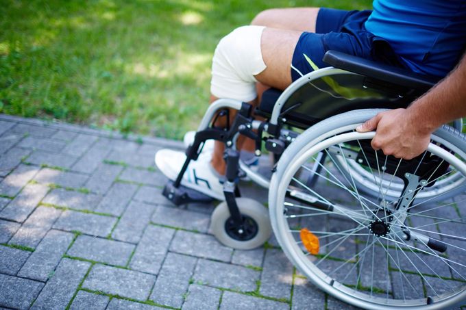 Una persona in sedia a rotelle, su un sentiero lastricato accanto a un prato, tiene in mano la ruota.