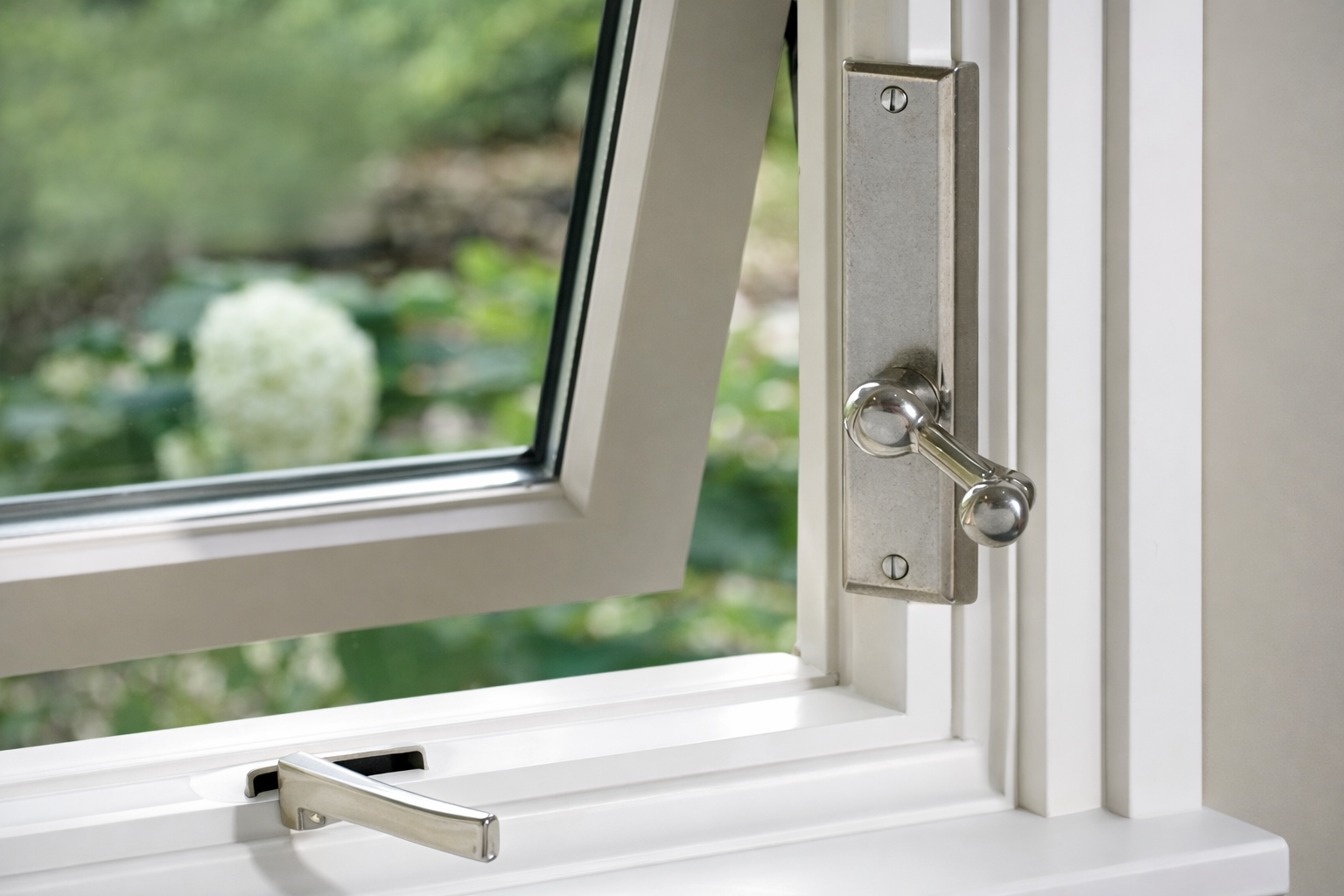 A close-up of a white window frame with a metal latch handle and a crank opener, partially open to a blurred garden view.