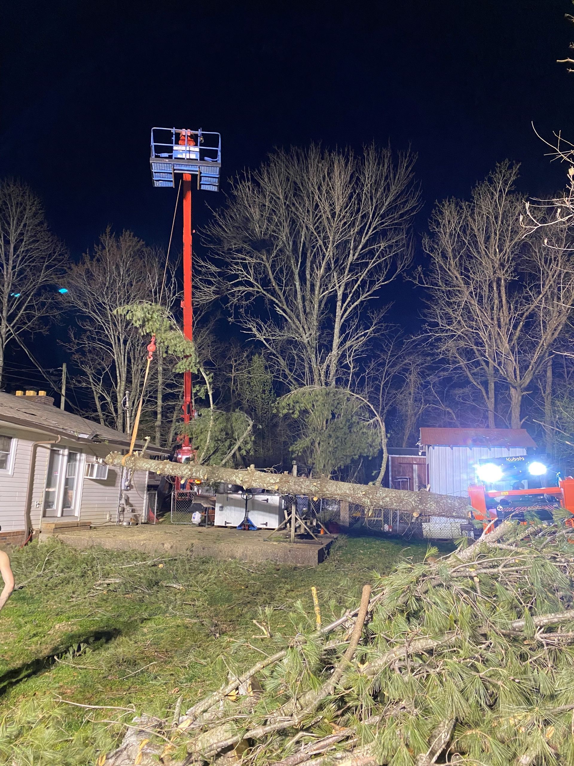 A tall red lift is raised near a house, with a tree being trimmed at night.
