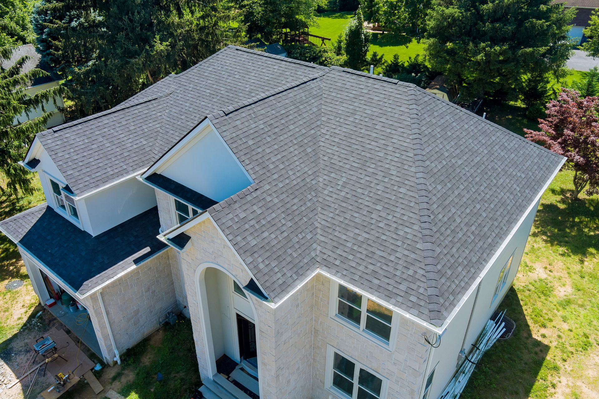 Aerial view of a two-story house with a gray shingled roof, beige siding, and surrounding trees.